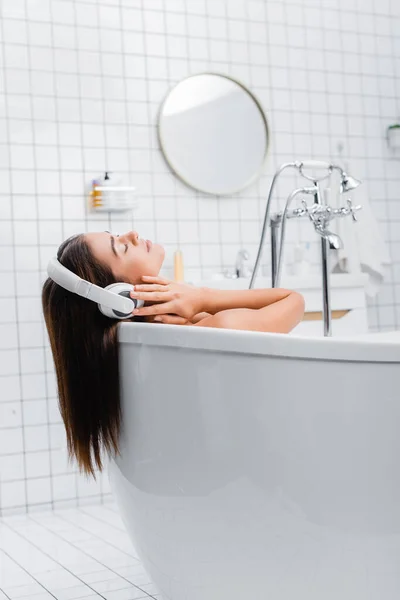 Mujer joven tomando baño y escuchando música en auriculares inalámbricos con los ojos cerrados - foto de stock