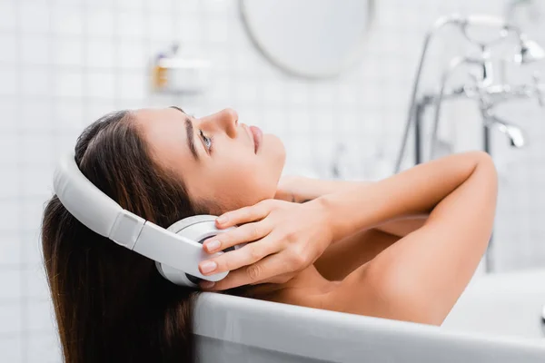 Mujer joven tocando auriculares inalámbricos mientras toma baño y escucha música - foto de stock