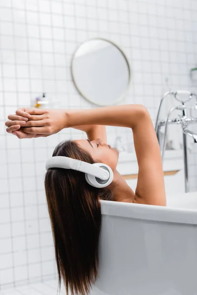 Mujer joven tomando baño y escuchando música en auriculares inalámbricos con los ojos cerrados - foto de stock