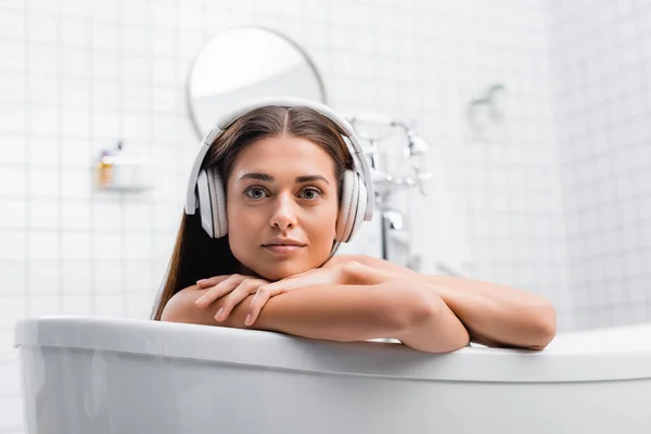 Jeune femme écouter de la musique dans les écouteurs sans fil tout en prenant un bain et en regardant la caméra — Photo de stock