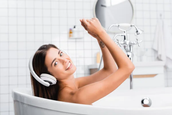 Веселая женщина улыбается в камеру, принимая ванну и слушая музыку в беспроводных наушниках — стоковое фото