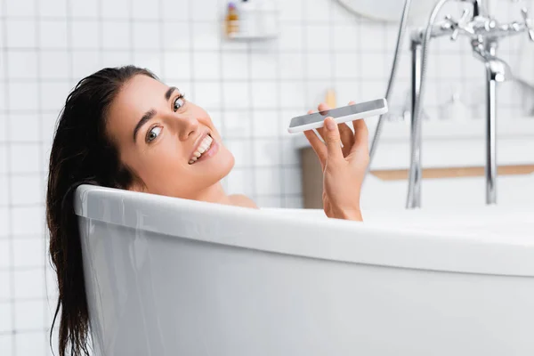 Happy young woman looking at camera while taking bath and talking on mobile phone — Stock Photo