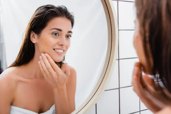 Lächelnde Frau, die im Spiegel schaut, während sie im Badezimmer das Gesicht schrubbt, verschwommener Vordergrund — Stockfoto