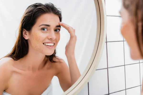 Happy woman applying scrub on face near mirror in bathroom, blurred front ground — стоковое фото
