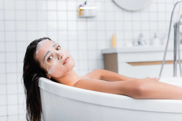 Junge Frau mit Gesichtsmaske entspannt sich in Badewanne und blickt in die Kamera — Stockfoto