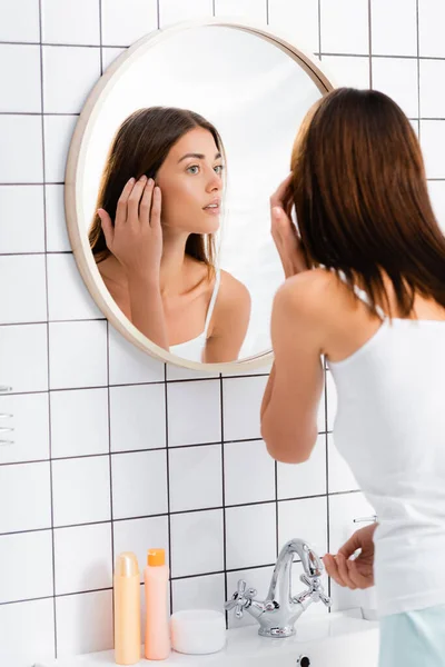 Young woman in white singlet looking in mirror in bathroom — Stock Photo