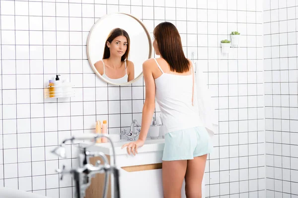 Joven mujer sonriente en soltera blanca y pantalones cortos de pie cerca del espejo en el baño, borrosa primer plano - foto de stock