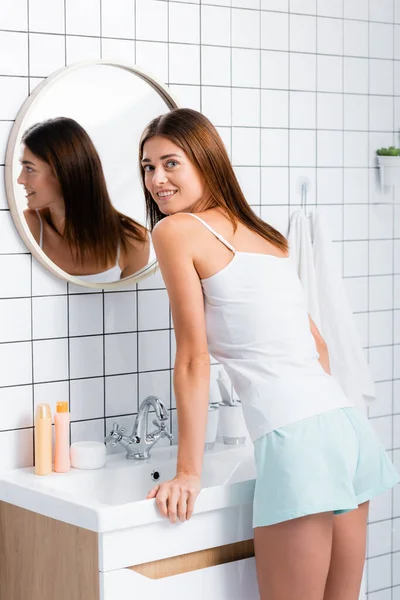 Feliz jovem mulher em shorts brancos e singlet sorrindo para a câmera enquanto de pé perto do espelho — Fotografia de Stock
