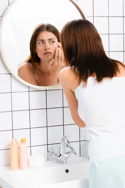 Unzufriedene junge Frau berührt Gesicht, während sie neben Spiegel im Badezimmer steht — Stockfoto