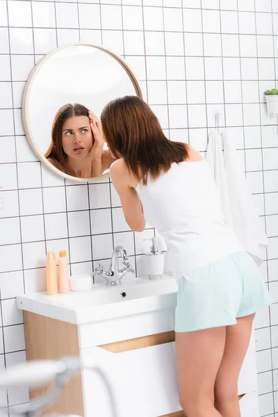 Unzufriedene Frau in weißen Shorts und Singlet berühren Gesicht, während sie neben Spiegel im Badezimmer stehen, verschwommener Vordergrund — Stockfoto