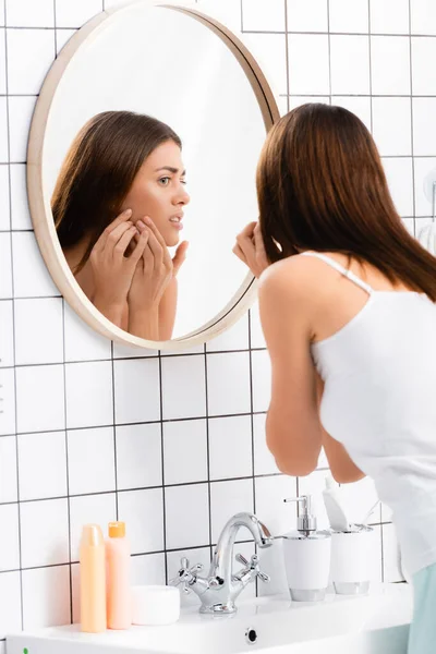 Unzufriedene Frau in weißem Singlet berührt Gesicht, während sie im Badezimmer in den Spiegel schaut — Stockfoto