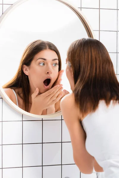 Conmocionada joven con la boca abierta tocando la cara mientras se mira en el espejo - foto de stock