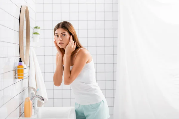 Besorgte junge Frau in Unterhemd und kurzer Hose berührt Gesicht, während sie im Badezimmer in die Kamera schaut — Stockfoto