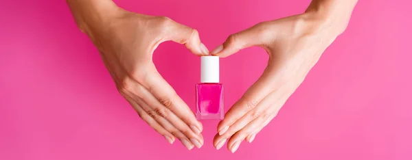 Vue partielle de la femme faisant signe de coeur avec les mains tout en tenant flacon d'émail d'ongle sur fond rose, bannière — Photo de stock