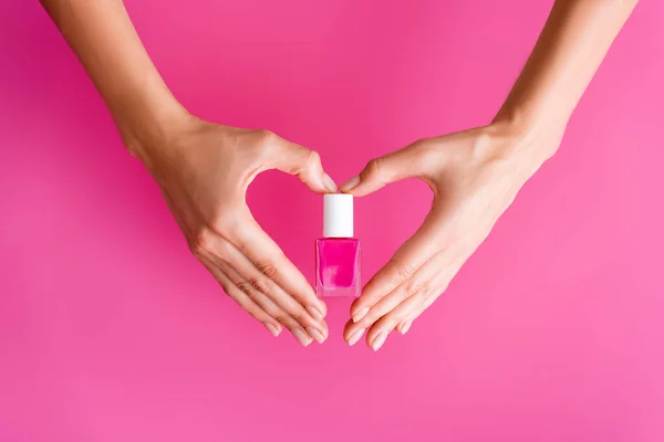 Vista cortada da mulher fazendo símbolo do coração com as mãos enquanto segurando garrafa de esmalte de unhas no fundo rosa — Fotografia de Stock