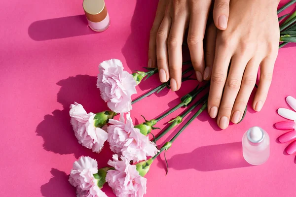 Top view of female hands near carnation flowers, palette of fake nails, vials with cuticle remover and nail varnish on pink background — Stock Photo