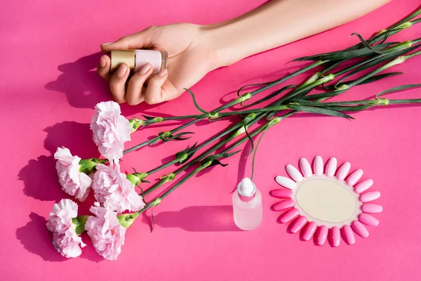 Vista recortada de la mujer sosteniendo esmalte de uñas pastel cerca de flores de clavel, removedor de cutículas y paleta de uñas artificiales sobre fondo rosa - foto de stock