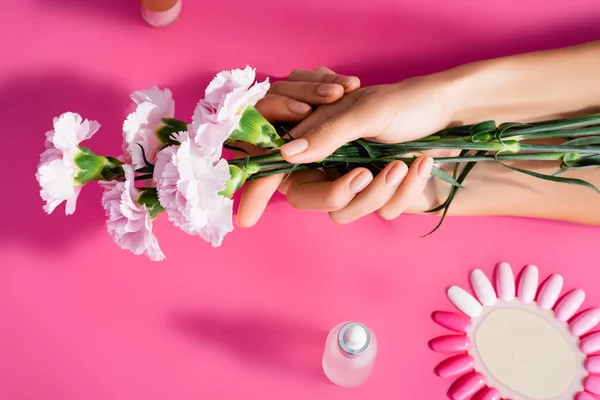 Vista recortada de la mujer sosteniendo flores de clavel cerca de la paleta de uñas artificiales y removedor de cutículas sobre fondo rosa - foto de stock