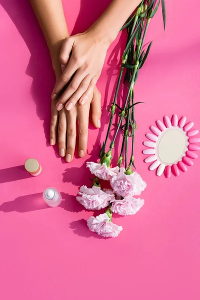 Top view of female hands near carnation flowers, palette of artificial nails, cuticle remover and nail polish on pink background — Stock Photo