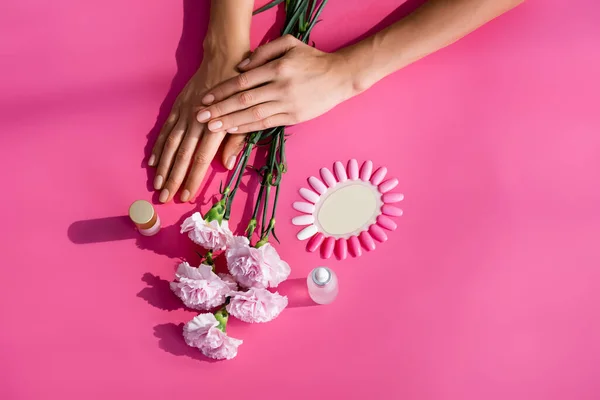 Top view of female hands near palette of false nails, carnation flowers, and bottles of cuticle remover and enamel on pink background — Stock Photo