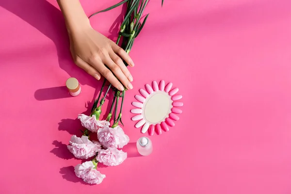 Вид зверху жіночих рук біля пляшок з лаком для нігтів та кутикули, квіти гвоздик та палітра підроблених нігтів на рожевому фоні — стокове фото