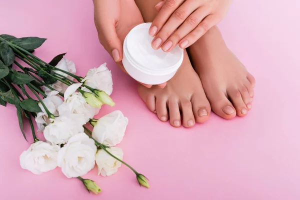 Visão parcial da mulher descalça segurando creme cosmético perto de flores eustoma branco no fundo rosa — Fotografia de Stock