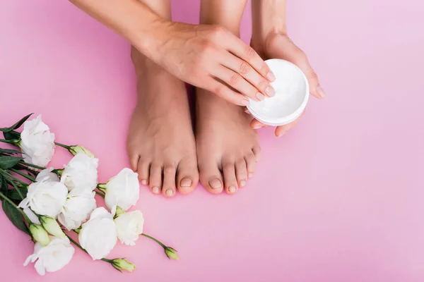 Vista cortada de mulher descalça segurando creme cosmético perto de flores eustoma branco no fundo rosa — Fotografia de Stock