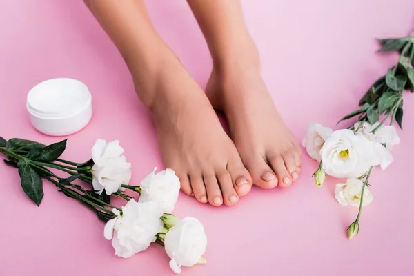 Vista superior de flores de eustoma cerca de crema cosmética y piernas femeninas con pedicura pastel sobre fondo rosa - foto de stock