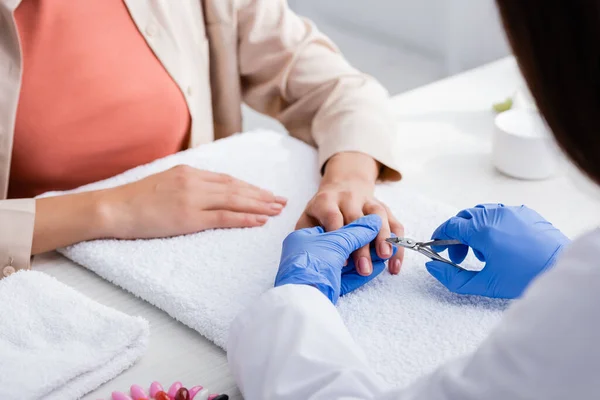 Partial view of manicurist cutting cuticle with nipper while making manicure to client, blurred foreground — Stock Photo