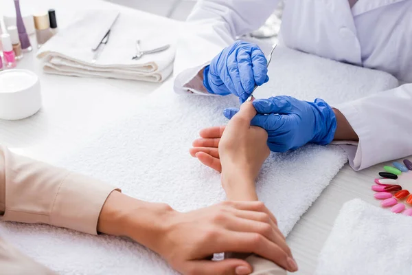 Partial view of manicurist in latex gloves using cuticle pusher while making manicure to client — Stock Photo