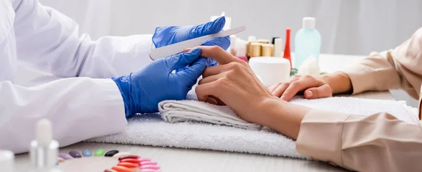 Vista parziale della manicure che tiene la lima per unghie mentre fa manicure al cliente in primo piano sfocato, banner — Foto stock