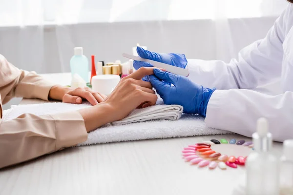 Vista recortada de la manicurista sosteniendo la lima de uñas mientras hace la manicura al cliente en primer plano borroso - foto de stock