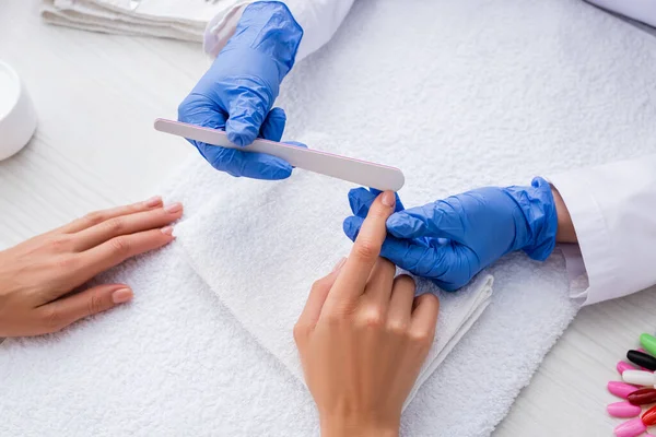 Partial view of manicurist in latex gloves using nail file while making manicure to woman — Stock Photo