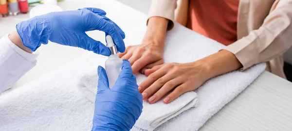 Vista recortada de manicurista sosteniendo botella de removedor de cutículas cerca del cliente, banner - foto de stock