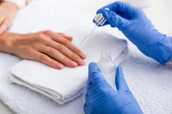 Vista parcial de la manicurista en guantes de látex aplicando removedor de cutículas en el dedo del cliente, fondo borroso - foto de stock