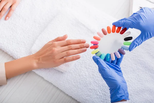 Vista parcial de la manicura sosteniendo muestras de uñas artificiales de colores cerca de la mano del cliente - foto de stock