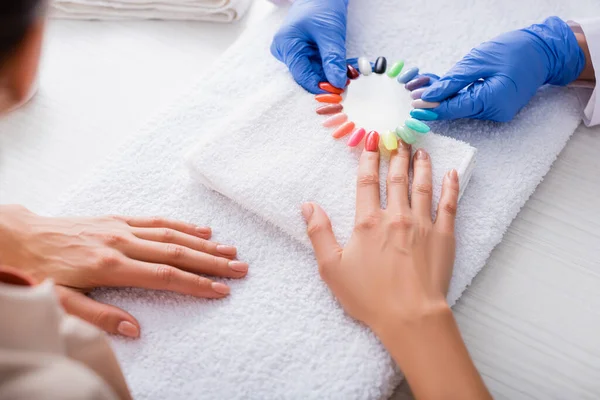 Partial view of manicurist in latex gloves holding palette of false nails near hand of woman — Stock Photo