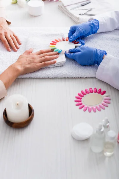 Cropped view of manicurist holding set of artificial nails near client and manicure supplies on blurred foreground — Stock Photo