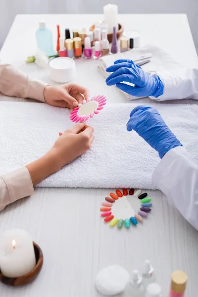 Vista cortada de mulher segurando paleta de unhas falsas perto manicure e manicure suprimentos no fundo borrado — Fotografia de Stock