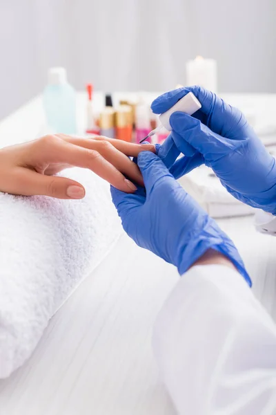 Vista recortada de la manicura en guantes de látex aplicando esmalte de uñas mientras se hace la manicura al cliente - foto de stock