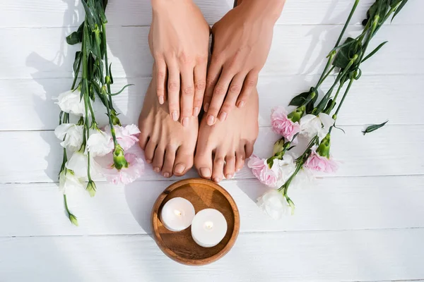 Vista superior de pies y manos femeninas con esmalte rosa pastel en clavos cerca de flores y velas en superficie de madera blanca - foto de stock