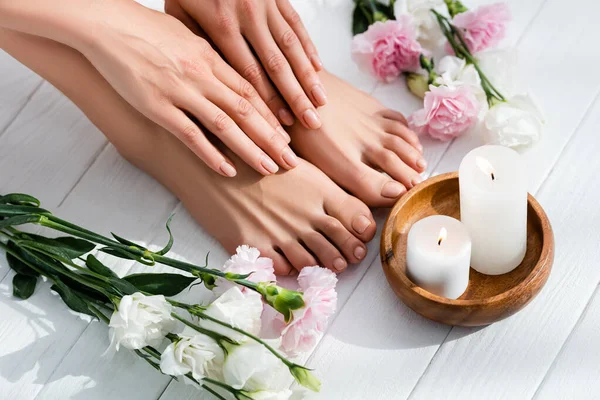 Weibliche Füße und Hände mit rosa glänzenden Nägeln in der Nähe von Nelken und Eustoma-Blumen und Kerzen auf weißer Holzoberfläche — Stockfoto