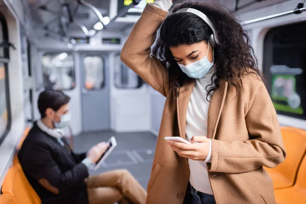 Joven afroamericana mujer en máscara médica escuchar música y el uso de teléfono inteligente cerca de hombre en el metro en fondo borroso - foto de stock