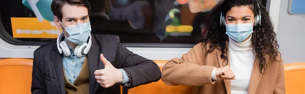 Pareja interracial en máscaras médicas golpeando codos en el metro, pancarta - foto de stock