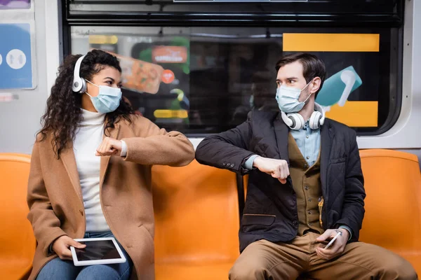 Coppia multiculturale in maschere mediche urtando gomiti e tenendo gadget in metropolitana — Foto stock