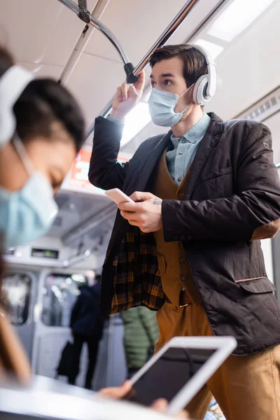 Man in headphones holding smartphone near african american woman in medical mask using digital tablet on blurred foreground — Stock Photo