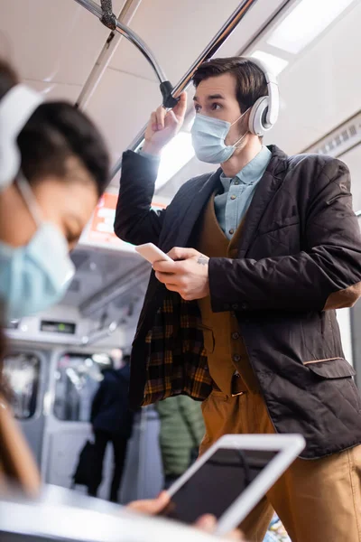 Mann mit Kopfhörer hält Smartphone in der Nähe afrikanisch-amerikanische Frau in medizinischer Maske mit digitalem Tablet auf verschwommenem Vordergrund — Stockfoto