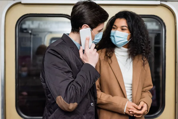 Mulher americana africana em máscara médica olhando para o namorado falando no smartphone no metrô — Fotografia de Stock