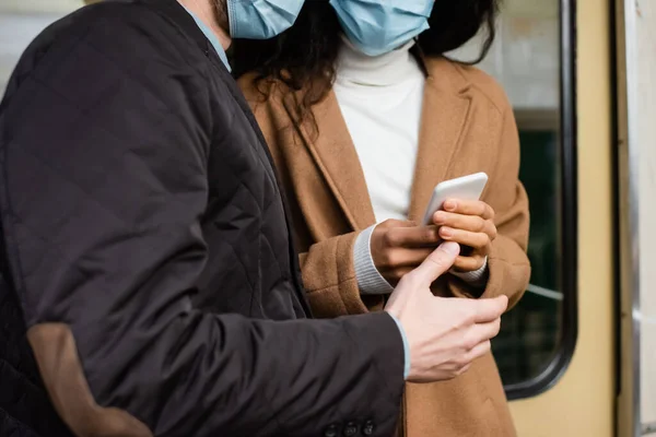 Vista recortada de la mujer afroamericana en máscara médica usando teléfono inteligente cerca de hombre en metro - foto de stock