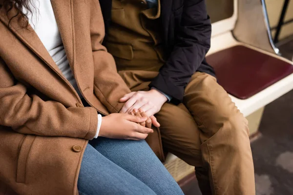 Vista cortada de casal interracial de mãos dadas enquanto sentado no vagão do metrô — Fotografia de Stock